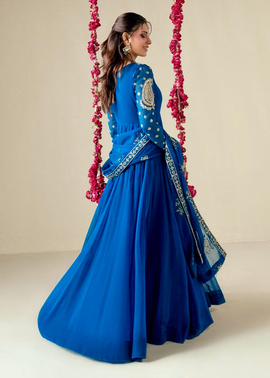 Woman in a blue traditional outfit with intricate designs, standing against a neutral background.