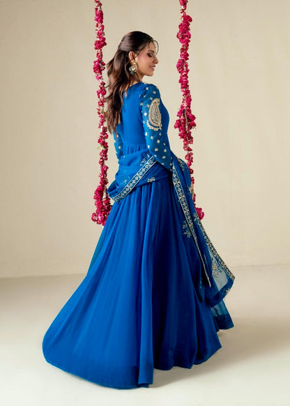 Woman in a blue traditional outfit with intricate designs, standing against a neutral background.