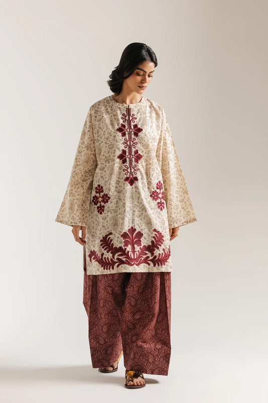 Woman wearing a traditional outfit with embroidered top and maroon pants on a plain background