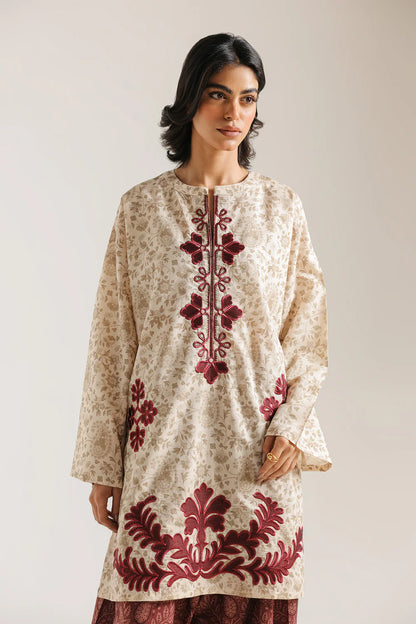 Woman wearing a cream-colored dress with red floral embroidery against a plain background