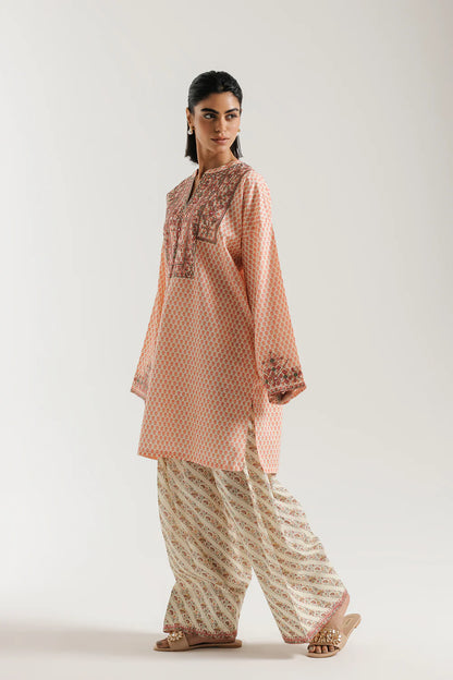 Woman wearing a peach-colored kurta with intricate embroidery and white pants on a light gray background
