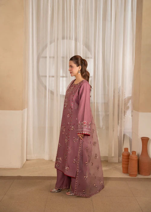 Woman in a pink embroidered traditional outfit standing in a room with white curtains and decorative vases.