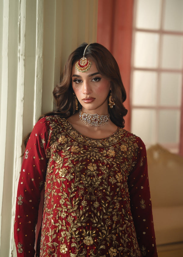 Woman in traditional red and gold embroidered outfit with jewelry, standing indoors.