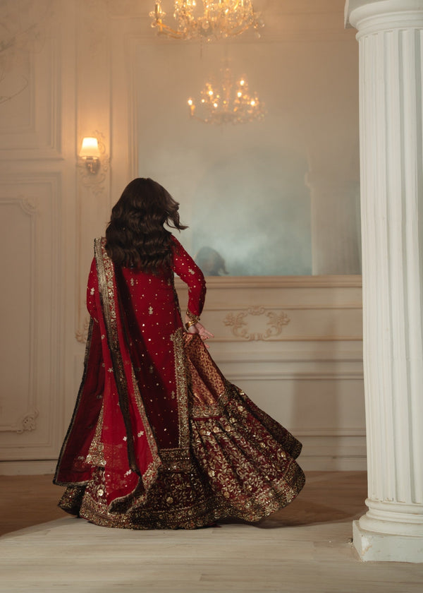 Person in a red and gold traditional outfit standing in a room with chandeliers and white walls.