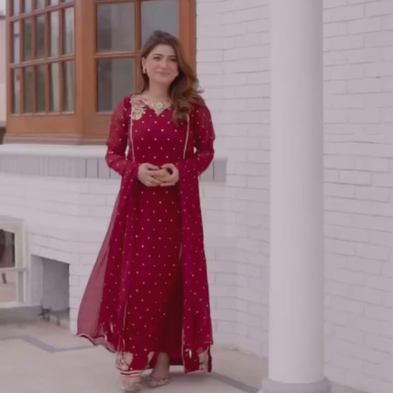 A model wearing an elegant deep red, gold-embroidered Pakistani designer lawn suit, showcasing the flowy fabric and sophisticated style in an outdoor setting