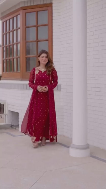 A model wearing an elegant deep red, gold-embroidered Pakistani designer lawn suit, showcasing the flowy fabric and sophisticated style in an outdoor setting